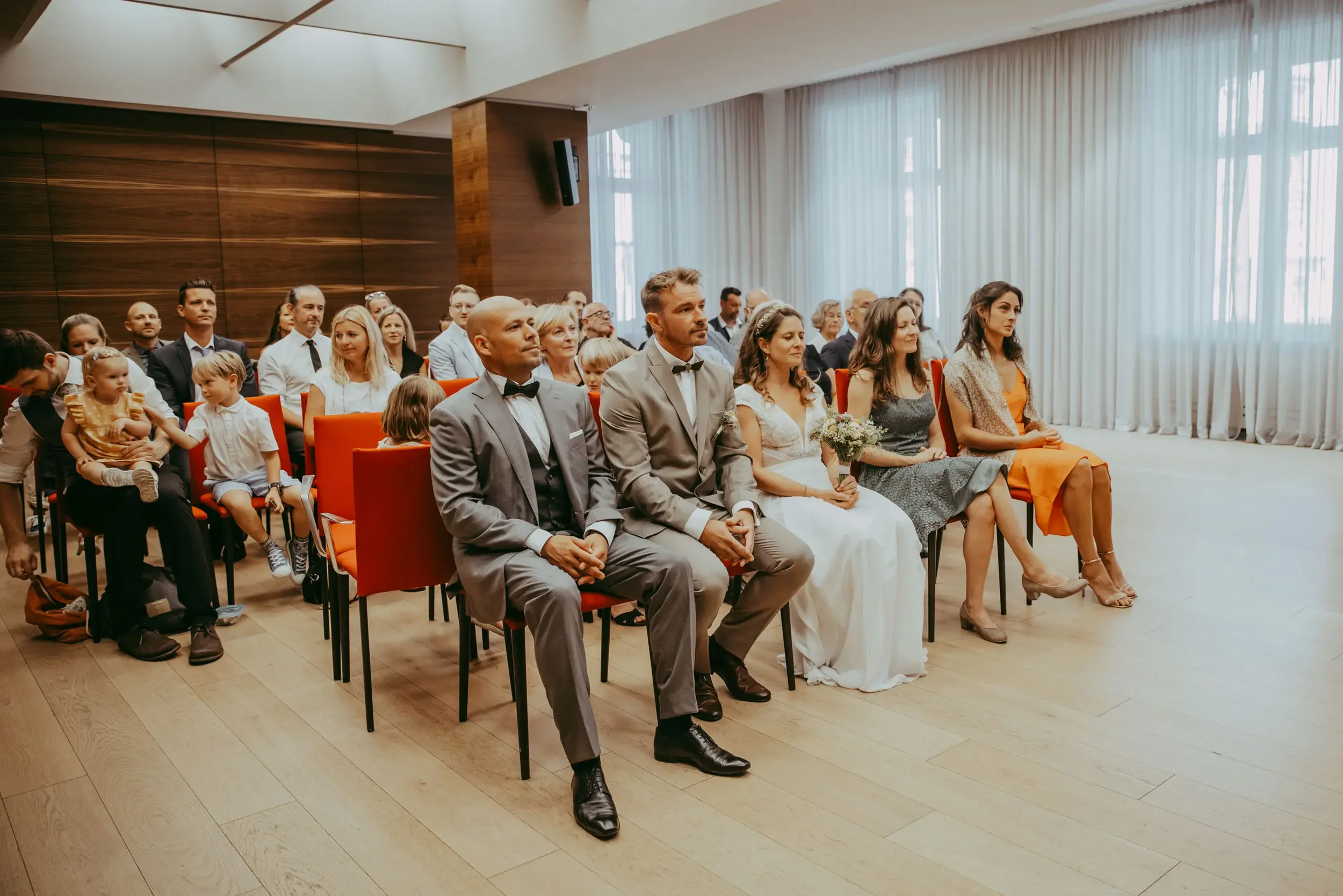 Hochzeitspaar mit Gästen im Saal bei einer Hochzeit, die vor einem großen Fenster sitzen.