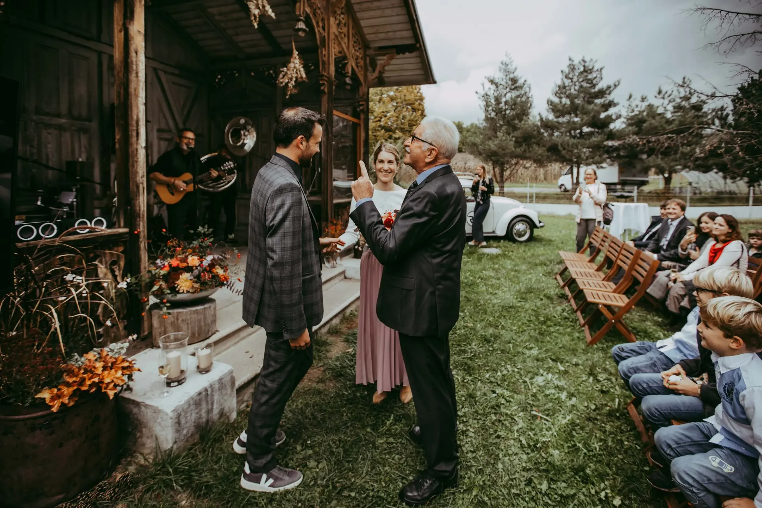 Ein älterer Mann und ein jüngerer Mann sprechen sich bei einer Hochzeitsfeier im Freien an. Im Hintergrund spielen Musiker auf einer kleinen Bühne. Gäste sitzen auf Bänken und freuen sich, während das Paar die Zeremonie auf einem Rasen im Freien voll