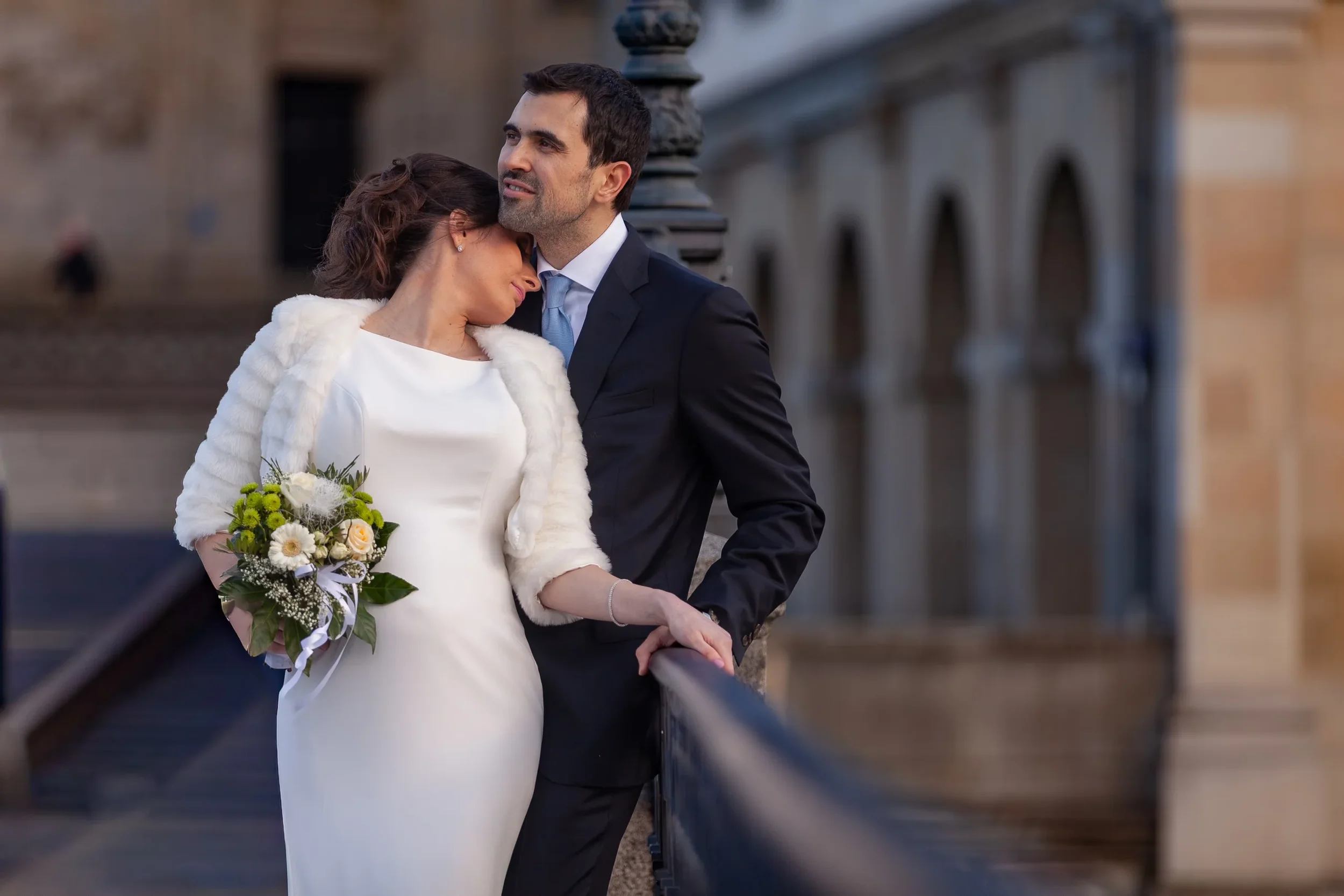 Ein Paar, vermutlich Braut und Bräutigam, steht eng beieinander an einer Brücke. Die Frau trägt ein weißes Hochzeitskleid und hält einen Blumenstrauß, der Mann trägt einen dunklen Anzug. Das Bild ist romantisch und emotional, mit einem historischen S
