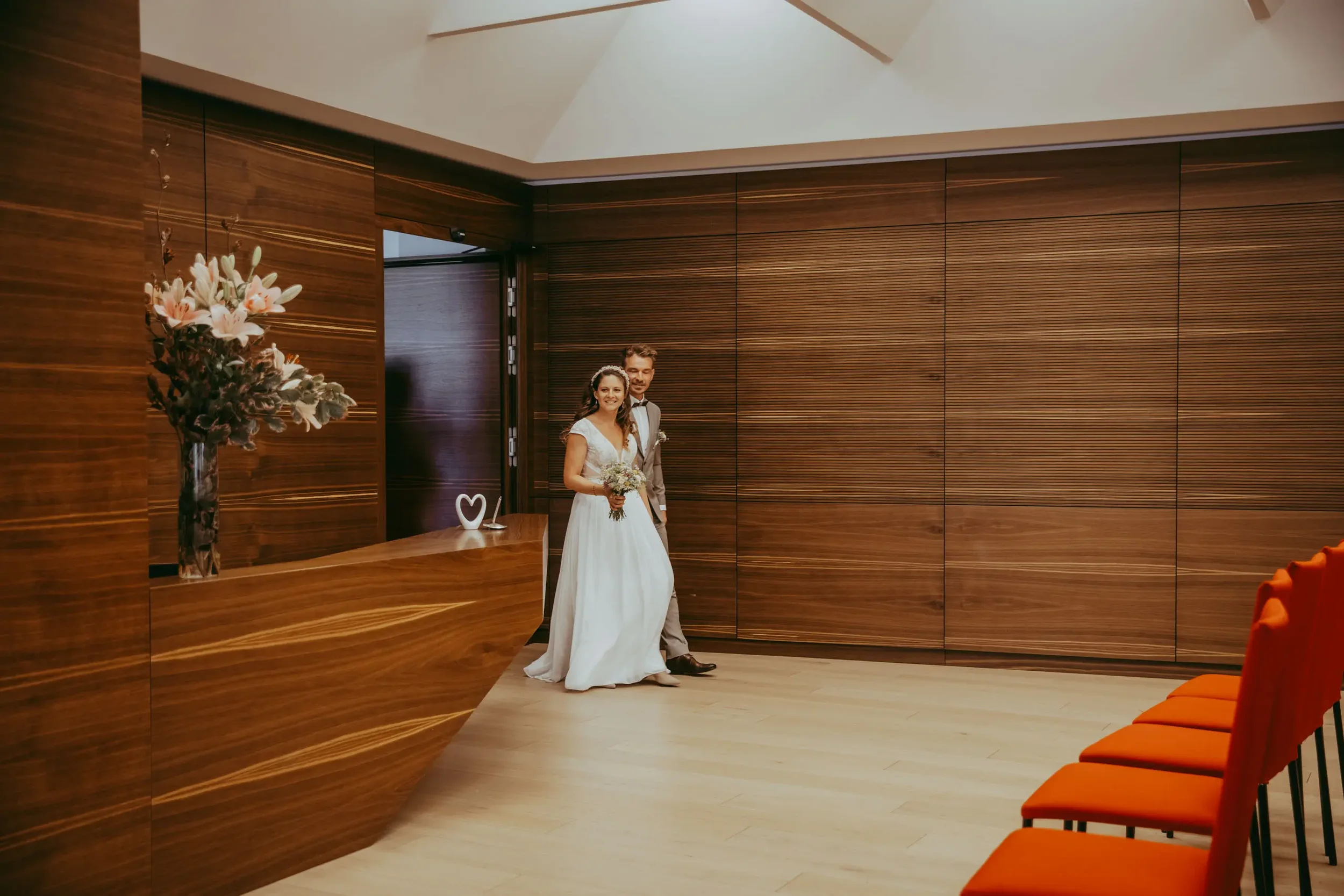 Ein Brautpaar steht in einem modernen Raum mit Holzvertäfelung und roten Stühlen an der Seite, die eine Hochzeit feiern.