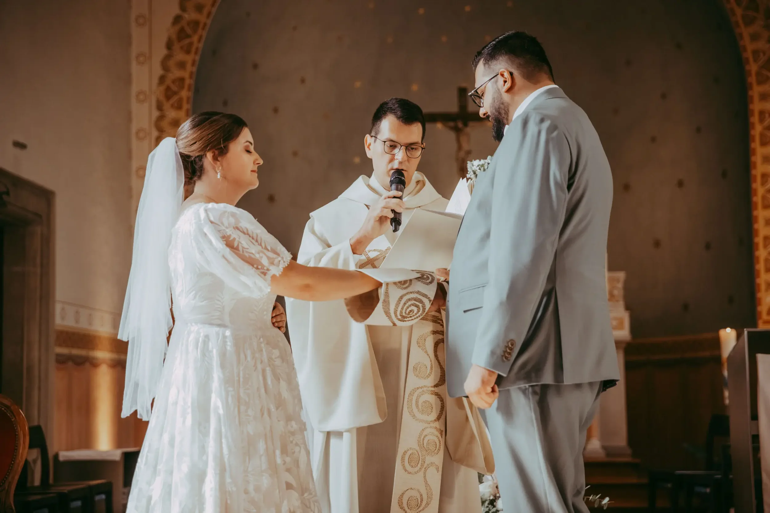 Ein Brautpaar bei ihrer Trauung in einer Kirche, mit einem Priester, der die Zeremonie vorliest, während der Bräutigam einen Handschein hält.