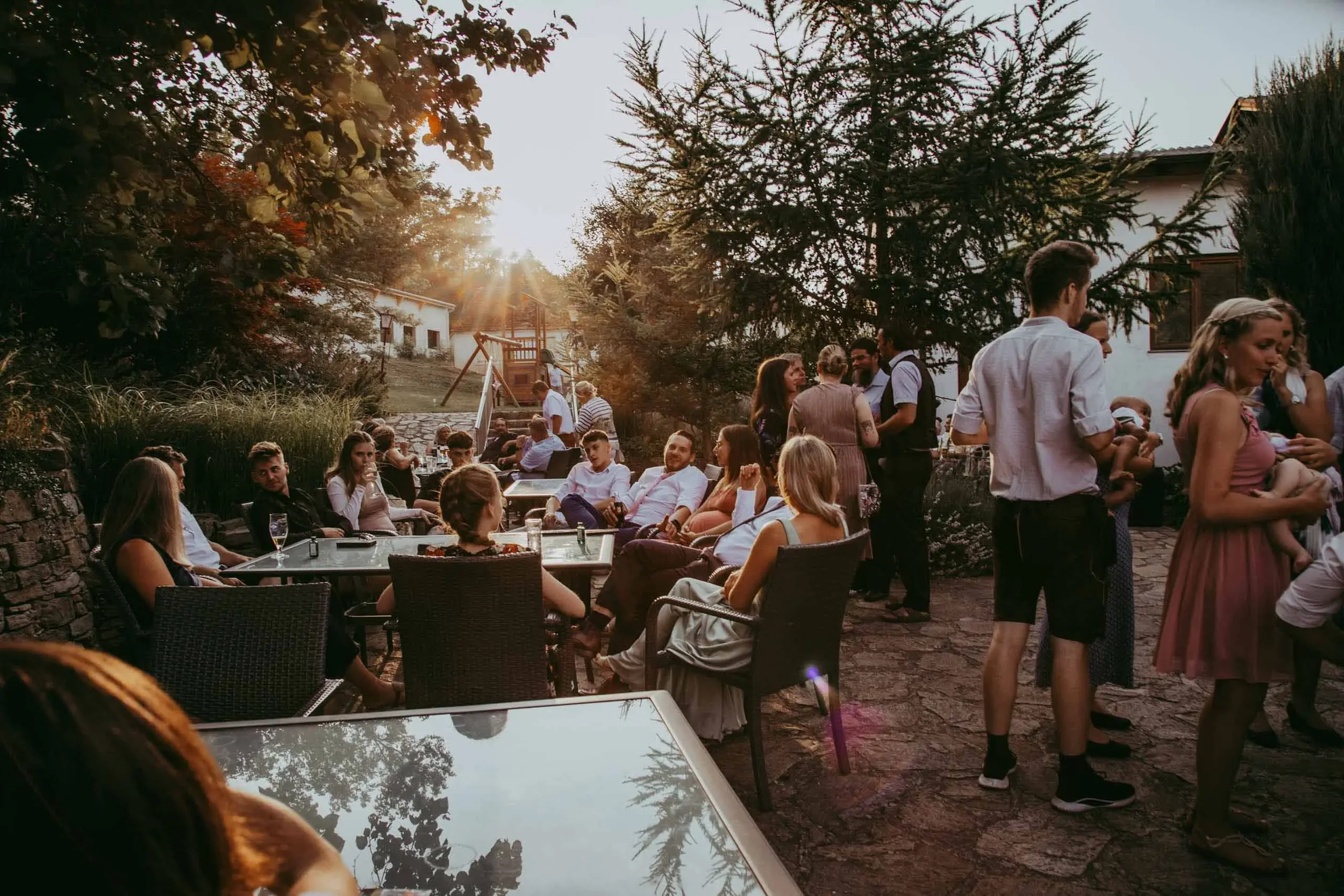 Eine Gruppe von Menschen versammelt sich im Freien, einige sitzen an Tischen, während andere stehen, bei Sonnenuntergang in einem Garten mit Bäumen und einem Spielplatz im Hintergrund.