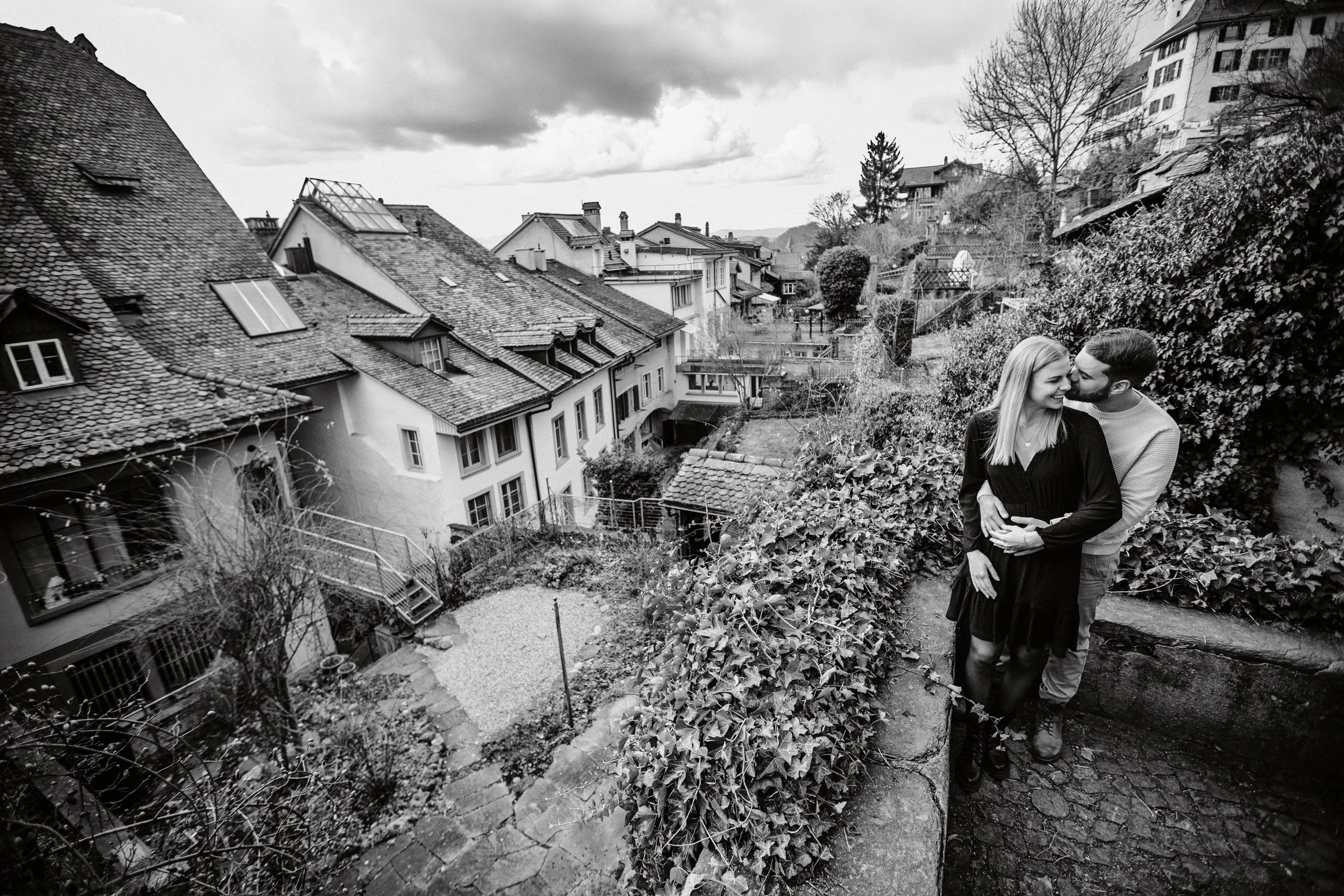 Ein lächelndes Paar, ein Mann und eine Frau, teilen sich einen Kuss auf einer Gartenmauer in einem Wohngebiet mit alten Dächern und modernen Gebäuden im Hintergrund. Es ist ein bewölkter Tag, und die Szene ist schwarz-weiß abgebildet.