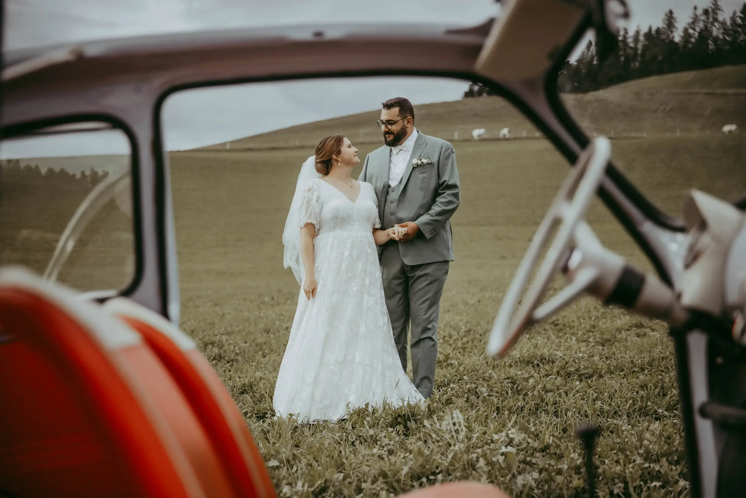 Ein Brautpaar, eine Frau in einem weißen Brautkleid und ein Mann in einem grauen Anzug, steht in einem offenen Feld, umgeben von Hügeln, während sie einander anschauen. Das Bild wird durch einen Rahmen aus roten und weißen Fahrrädern im Vordergrund e