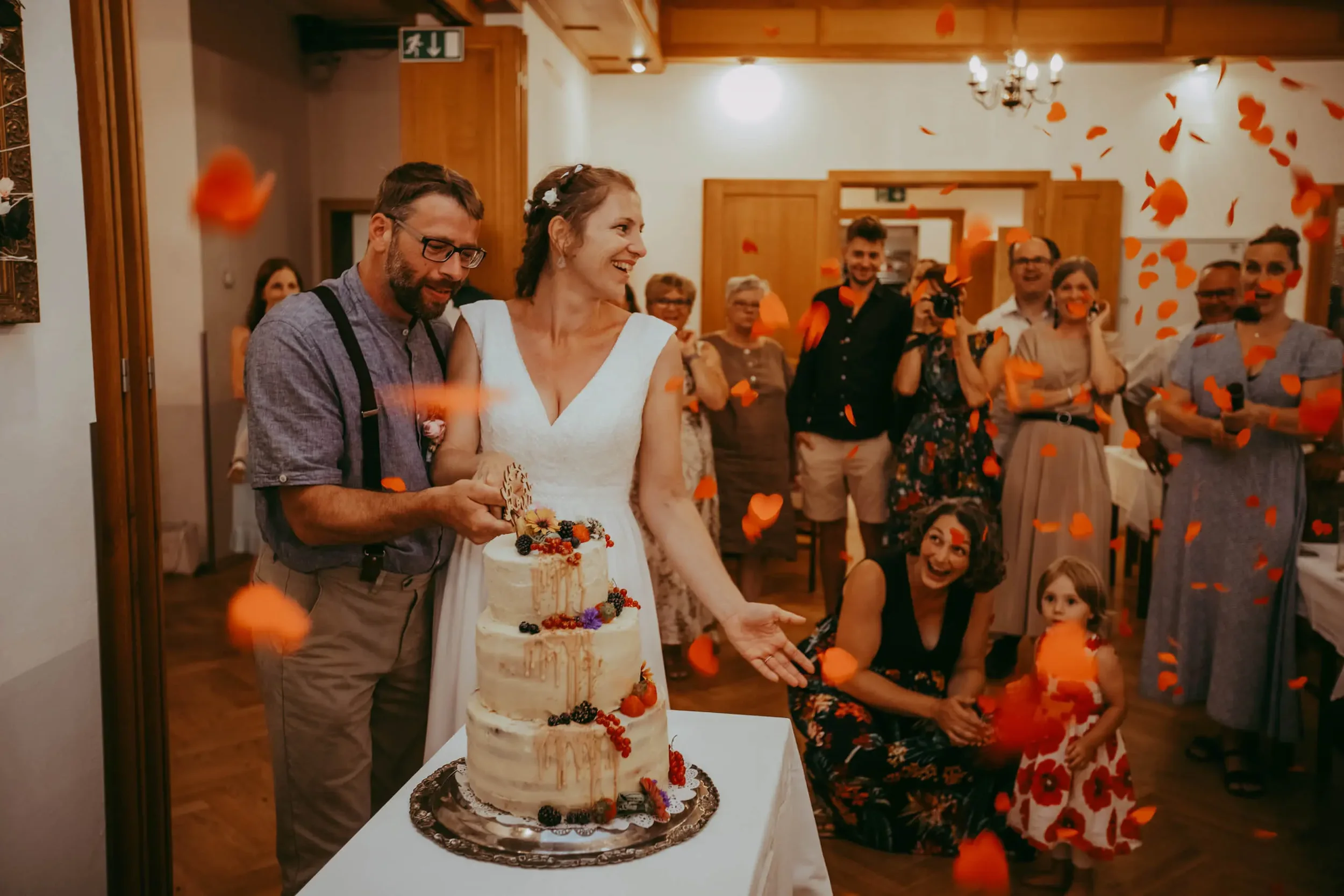 Ein Hochzeitspaar, das vor einer Gruppe von Gästen einen Hochzeitkuchen anschneidet, während orangefarbene Blätter im Raum herumschweben.