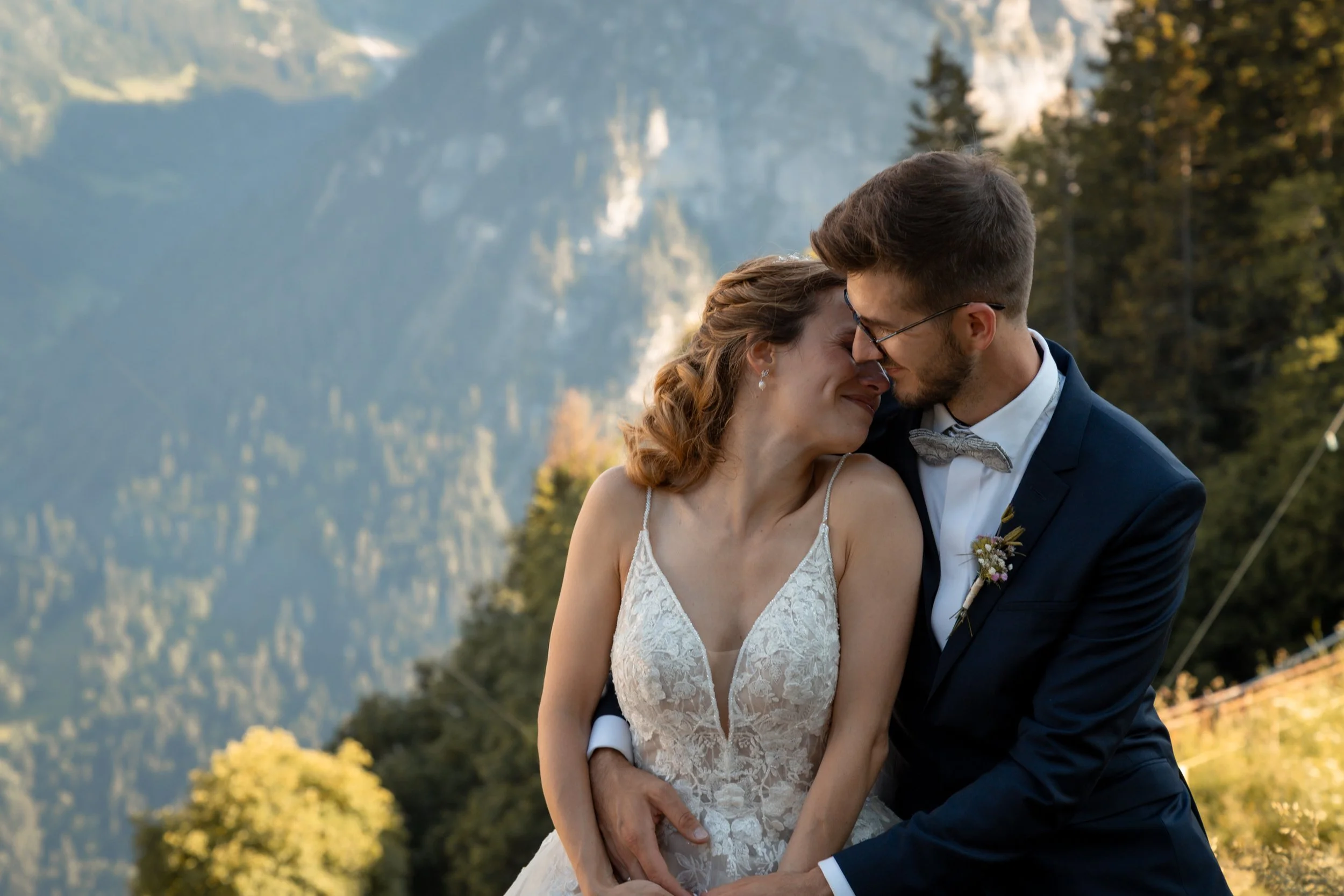 Ein Paar, vermutlich Braut und Bräutigam, steht sich mit geschlossenem Kopf und verschränkten Armen im Freien vor einer bergigen Landschaft. Die Frau trägt ein weißes Hochzeitskleid mit Spitze, der Mann einen dunklen Anzug mit Fliege.