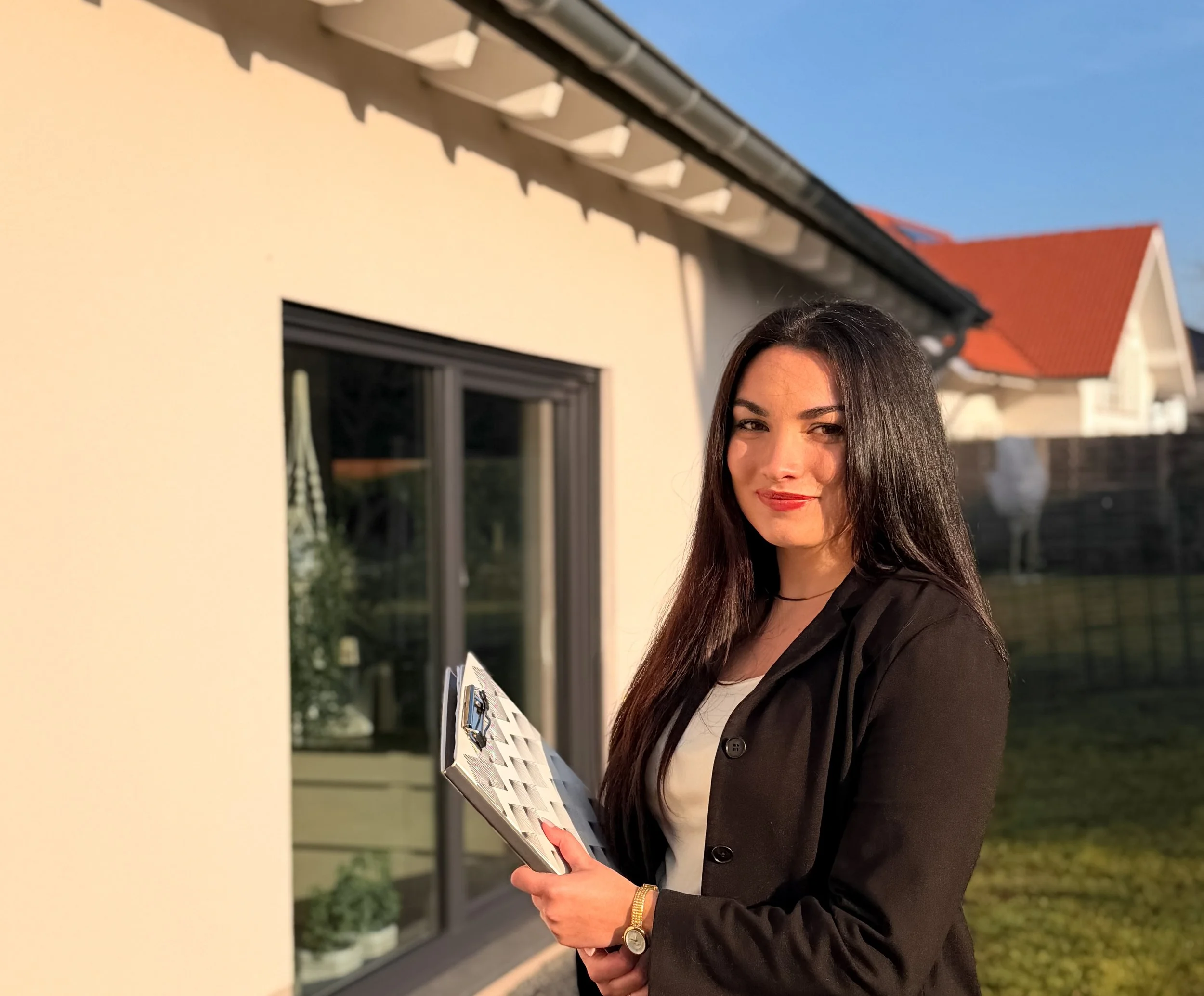 Eine junge Frau mit langen dunklen Haaren, Brille und dunklem Blazer hält eine Bewertungsbroschüre. Sie steht vor einem Haus mit großen Fenstern und rotem Dach, im Hintergrund ist blauer Himmel zu sehen.