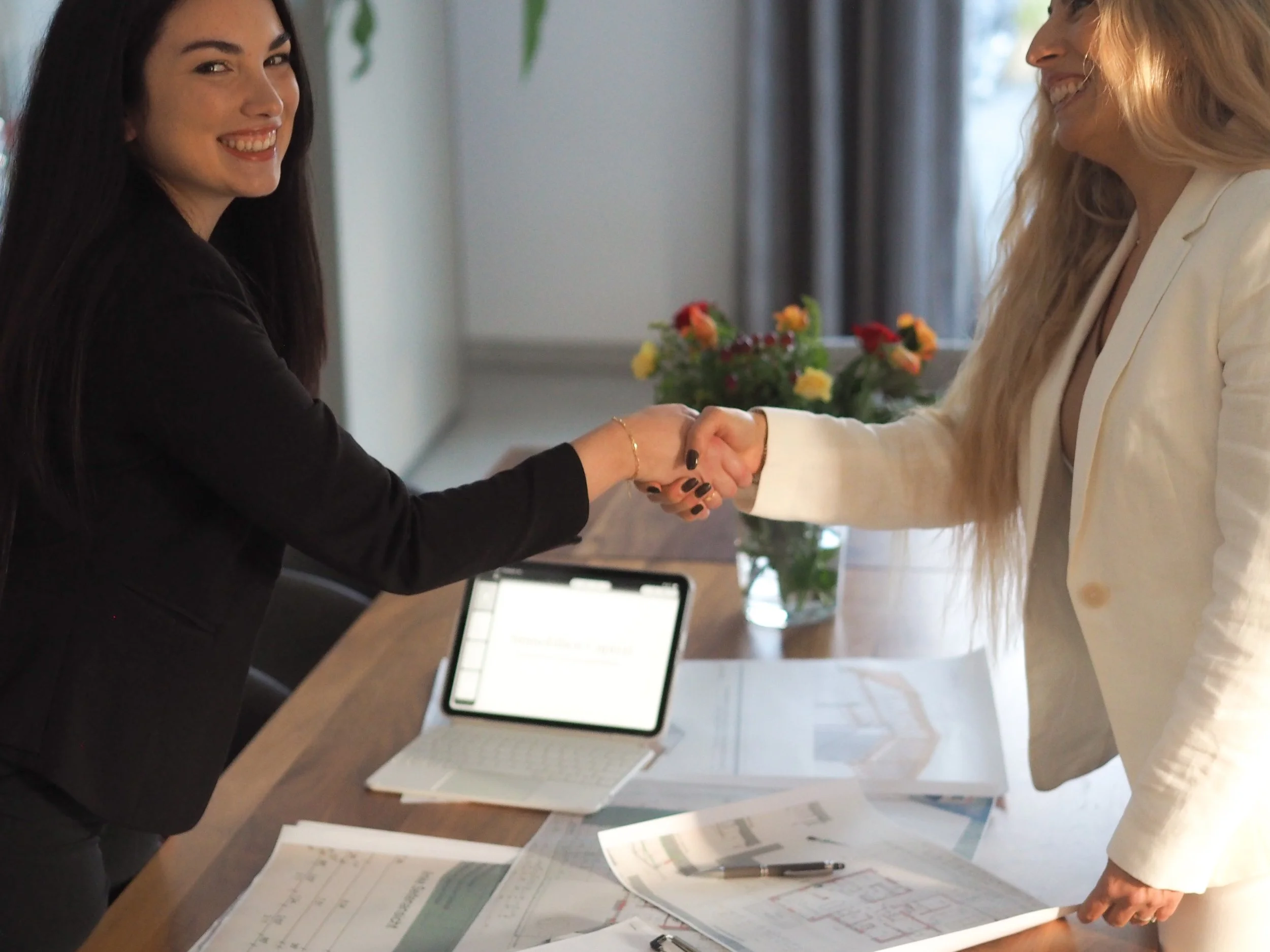 Giuseppina Capizzi und Carolina Gallace schütteln sich die Hände und lächeln sich bei einem Treffen im Büro an, mit einem Laptop und Dokumenten auf dem Tisch.