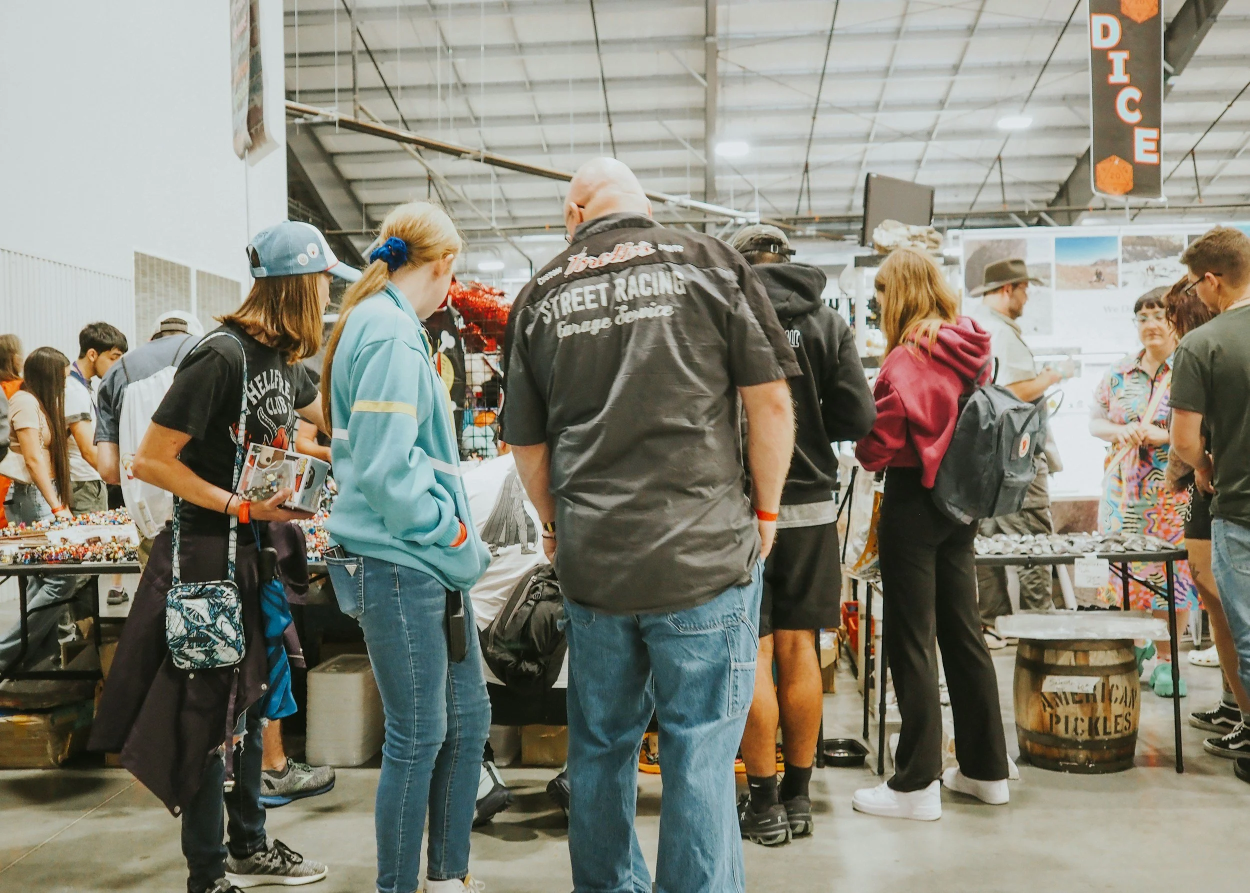 People shopping at a market or fair inside a large warehouse, with merchandise and signage visible.