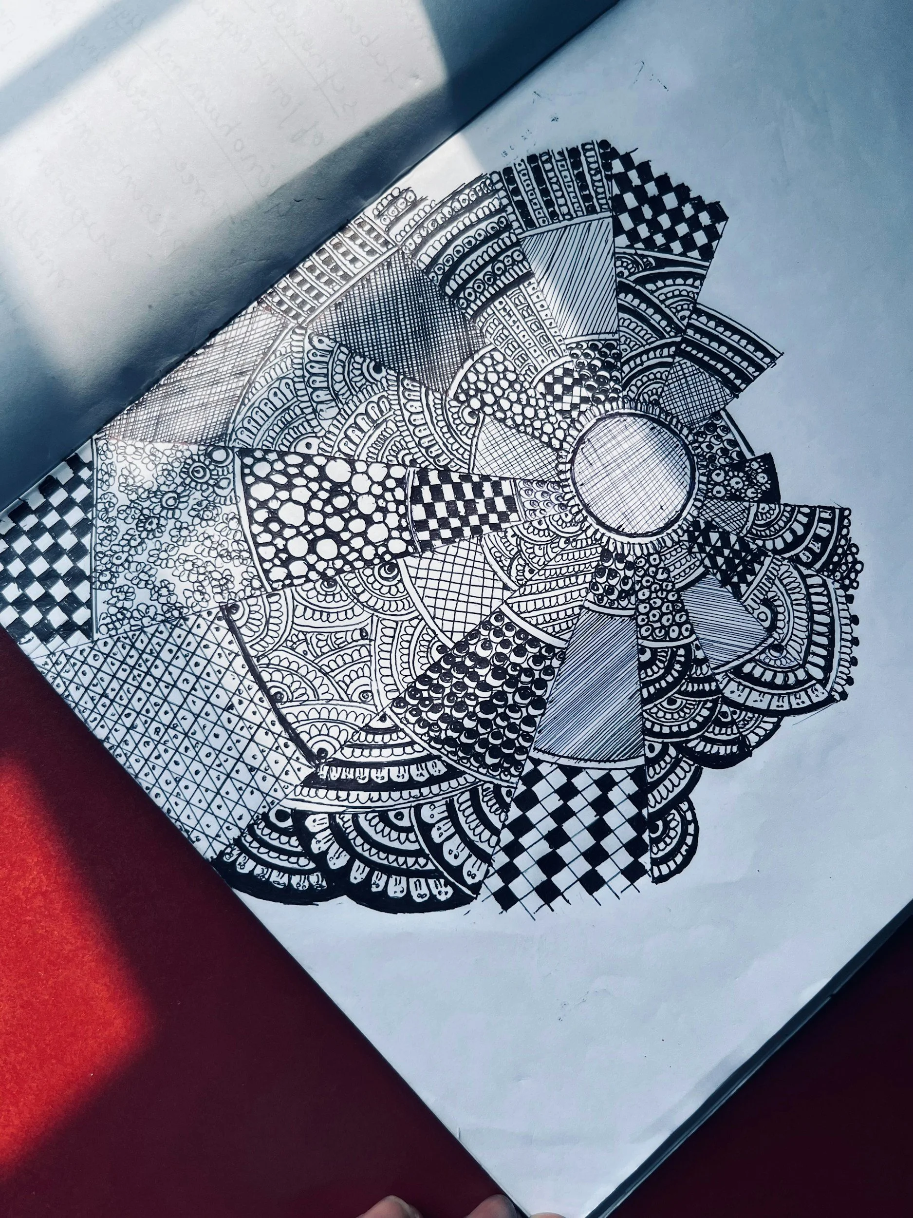 A black and white intricate mandala design drawn on white paper, featuring geometric shapes, patterns, and detailed line work.
