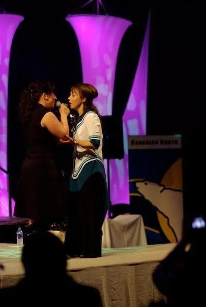 Two women stand on a stage, facing each other, holding a microphone and engaged in a conversation or performance, with purple lit stage lights in the background.