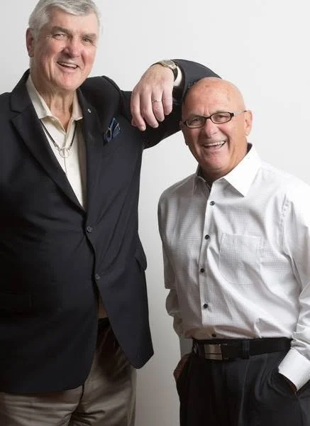 Two middle-aged men in formal business attire smiling, one resting his arm on the other's shoulder against a plain white background.