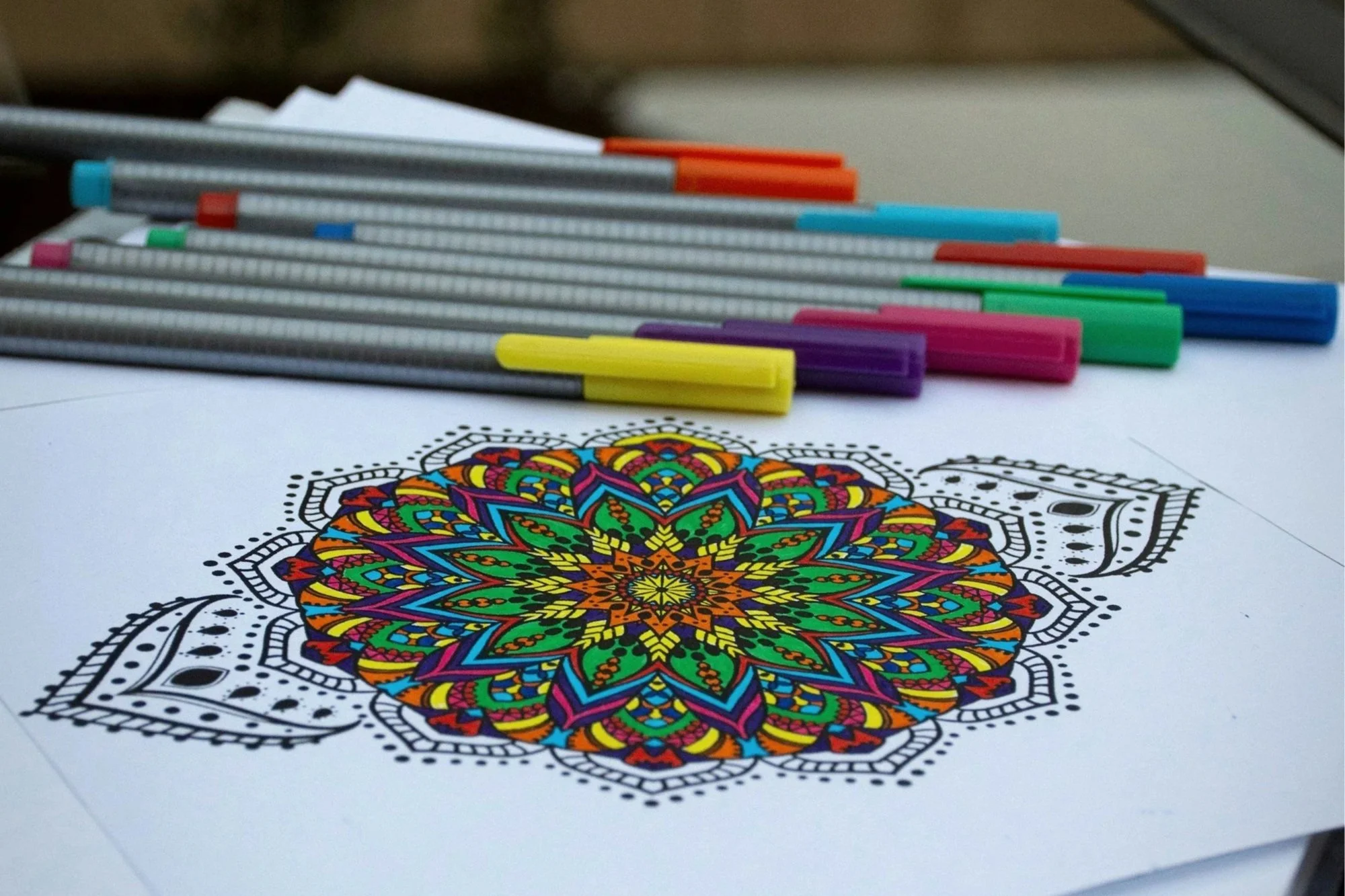 A colorful, intricate mandala coloring page on paper with multiple colored markers placed on top.