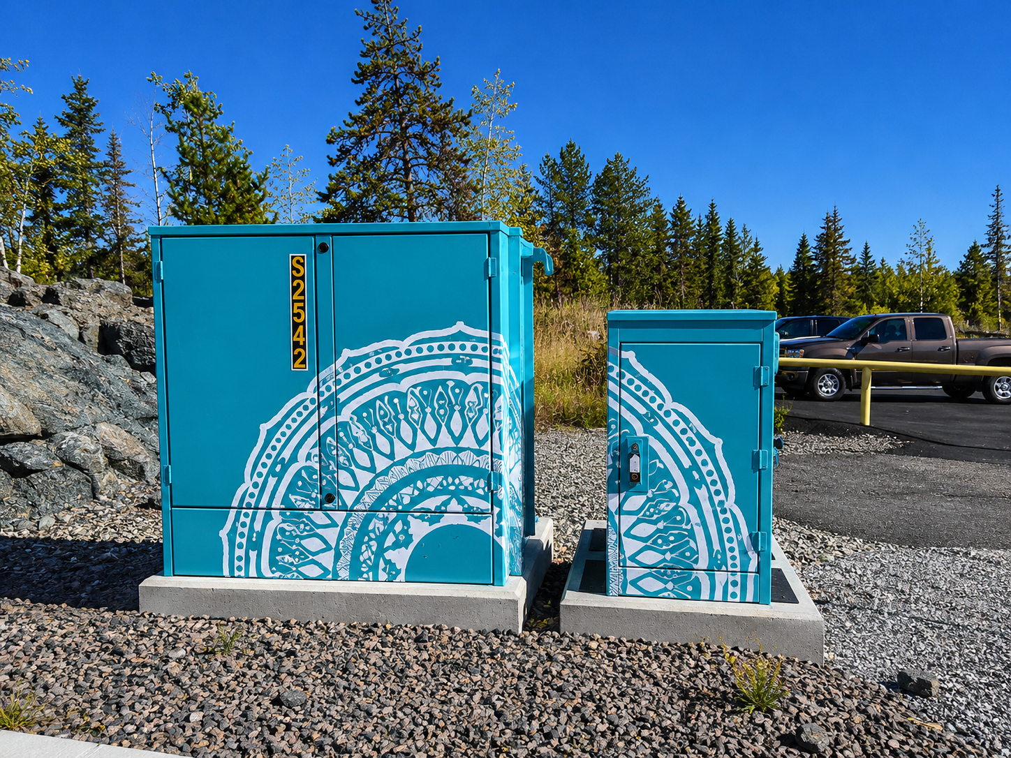 Electric utility boxes painted blue with white decorative patterns, situated on concrete pads in an outdoor parking area with trees and cars in the background.