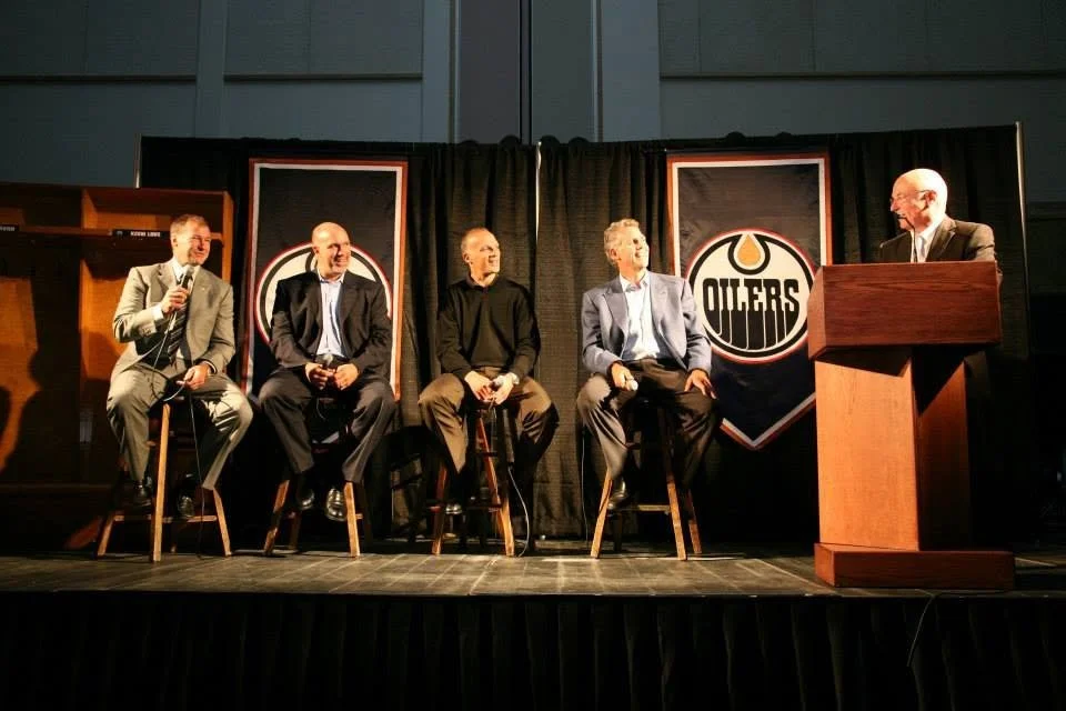 Five men are sitting on stools on a stage with black curtains and Edmonton Oilers logos behind them, participating in a panel discussion. One man is standing at a wooden podium.
