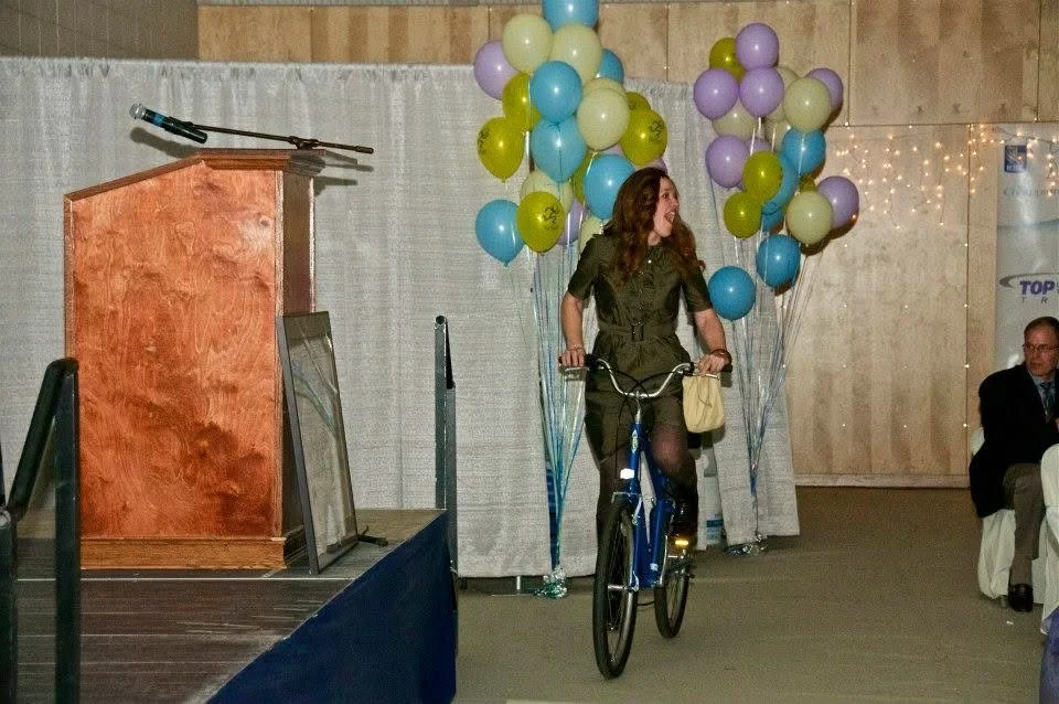 A woman riding a bicycle in front of a decorated backdrop with balloons at an indoor event.