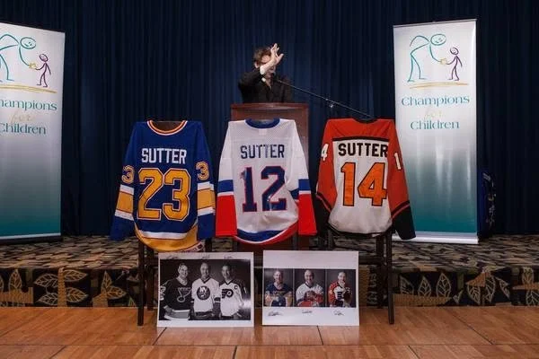 A person speaking at a podium during a charity event, with three hockey jerseys on display with the name Sutter and numbers 23, 12, and 14, and framed photographs of hockey players on a table in front of the jerseys.