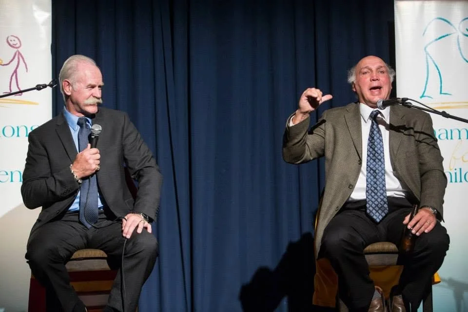 Two older men in business suits sitting on stage chairs, engaged in a conversation during a panel or interview, with blue curtains and a banner in the background.