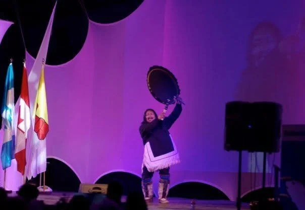 A woman on stage holding a drum above her head during a performance, with colorful flags to her left and a speaker to her right, under purple lighting.
