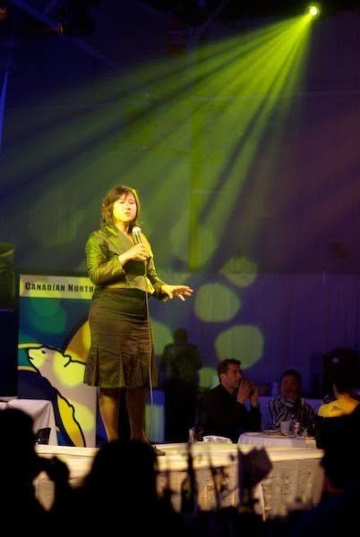 A woman standing on stage holding a microphone, speaking to an audience in a dimly lit room with yellow stage lights.
