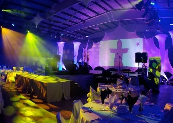 Event space with illuminated stage, decorated with nautical theme, featuring anchor projections and star designs, set up for a banquet with tables, chairs, and colorful lighting.