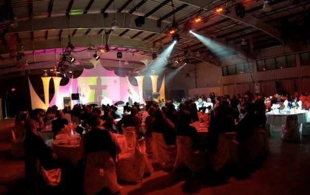 A large gathering of people seated at round tables at an indoor event, with stage lighting and a cross projected on the backdrop.