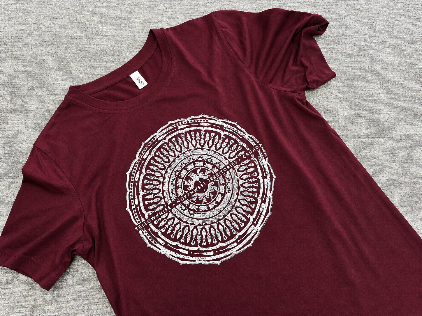 Maroon T-shirt with a white graphic of a circular emblem and text that reads "Yellowknife Northwest Territories Canada". The T-shirt is laid flat on a light-colored surface.