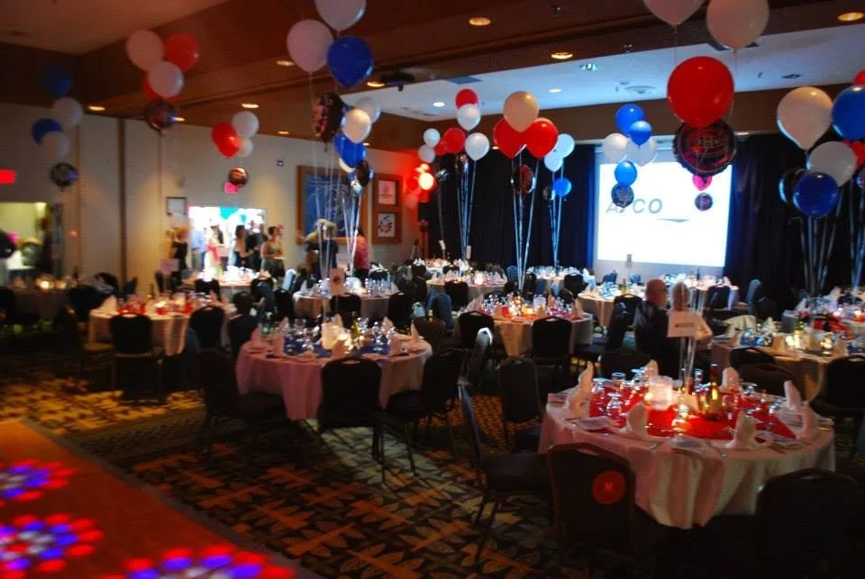 A decorated banquet hall set up for an event, with round tables covered in pink tablecloths, surrounded by chairs, and decorated with red, white, and blue balloons. A large screen displays the logo of Coca-Cola in the background.