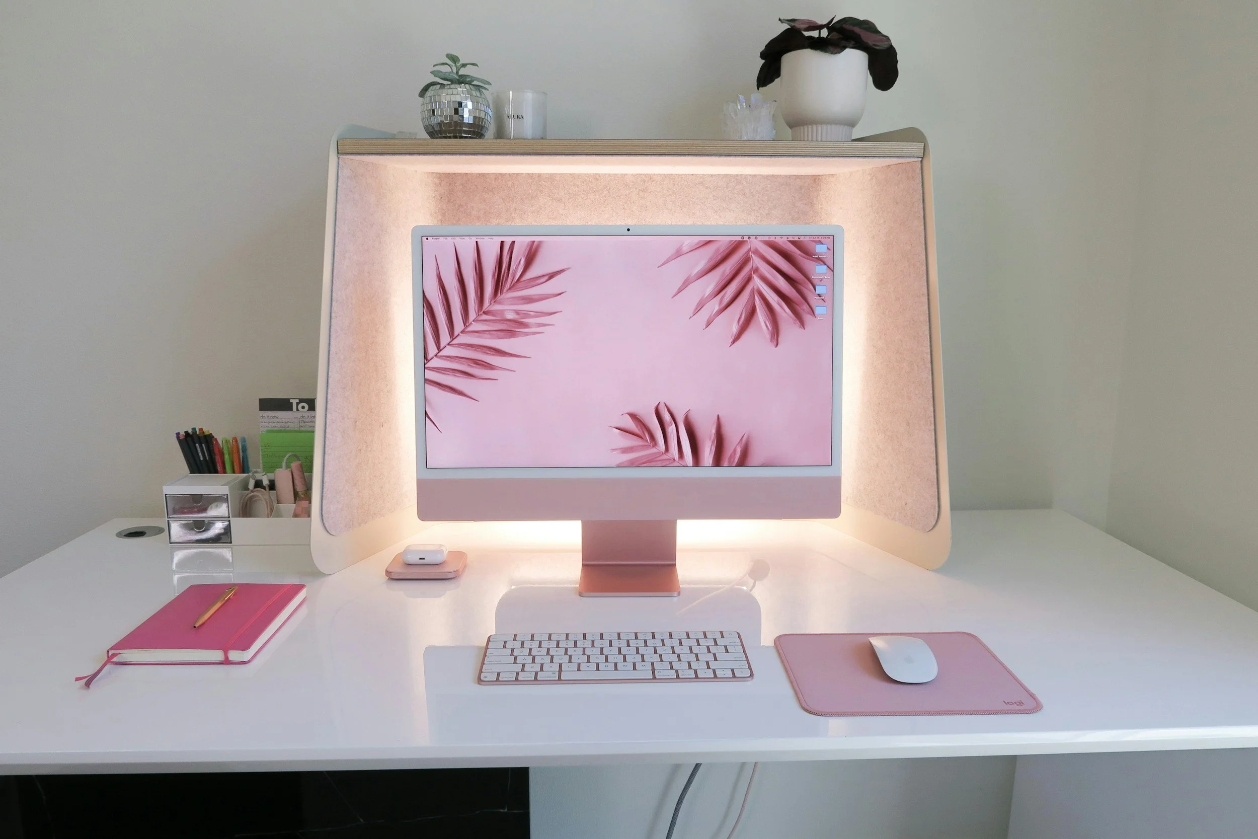 A pink-themed workspace with an iMac computer on a white desk, displaying a pink palm leaf background. The desk holds a pink notebook, a pink mouse pad with a mouse, a white wireless keyboard, and a closed case. To the left, there is a small white drawer unit with colored pens. On the top shelf, there are potted plants, candles, and decorative items. The background wall is light-colored.