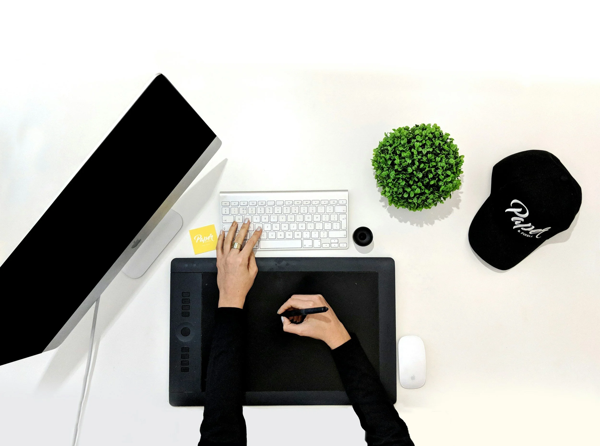 Overhead view of a white desk with a computer monitor, keyboard, and mouse, a potted green plant, a black cap, a small speaker, and two hands working on a drawing tablet, with a yellow paper labeled 'Paper'.