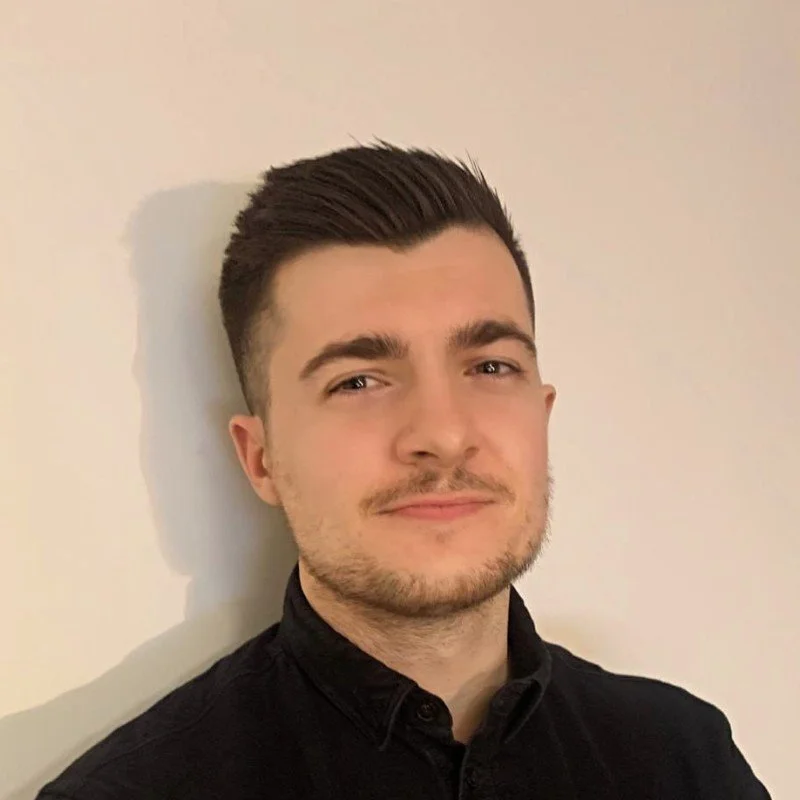 Simon Davies, independent business psychologist with short dark hair, light facial hair, wearing a black shirt, standing against a plain light-colored wall.