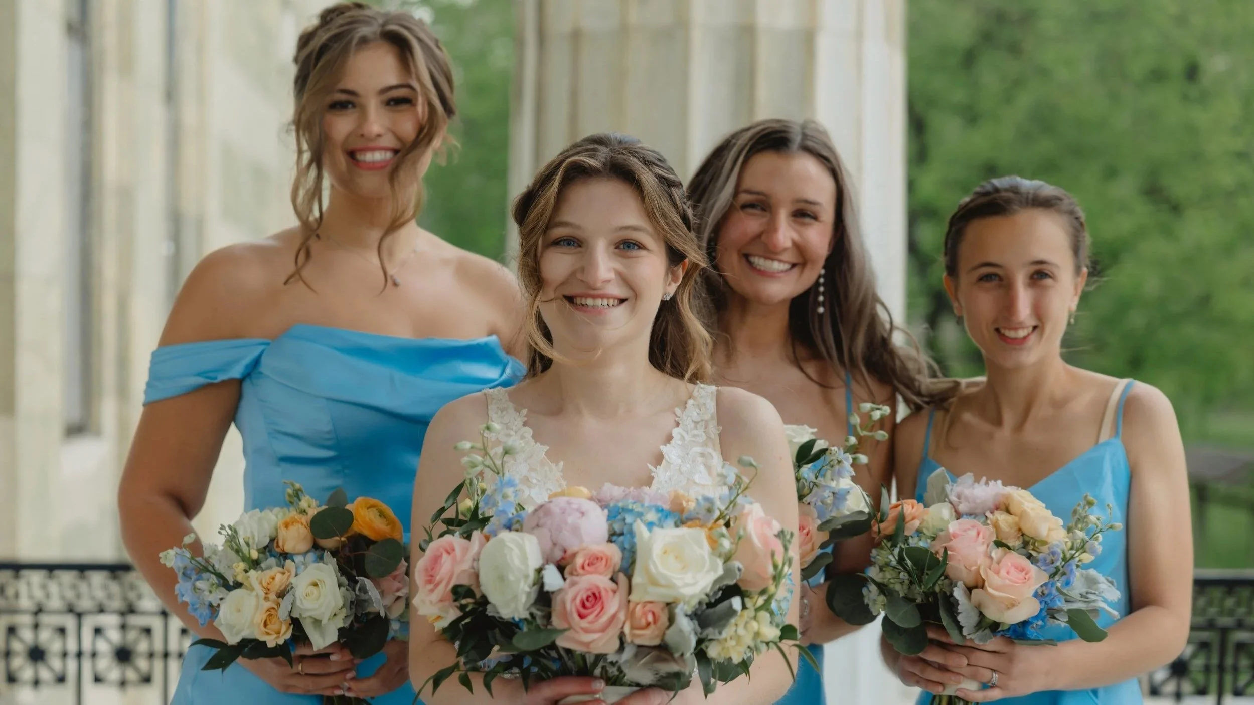 A group of four women smiling, holding floral bouquets, standing outdoors near a building with columns, with greenery in the background.