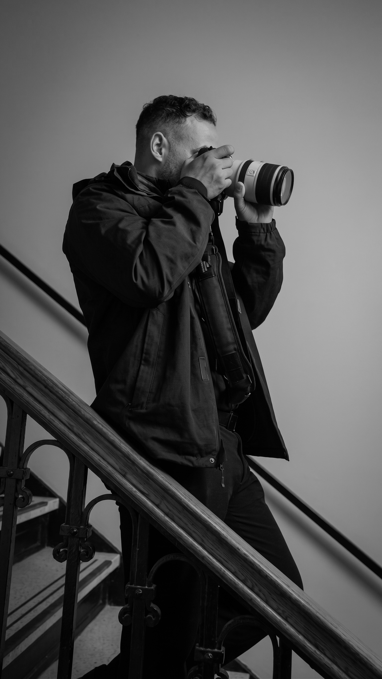 Black-and-white photo of a man with short curly hair taking a picture with a camera while standing on a staircase, wearing a dark jacket.