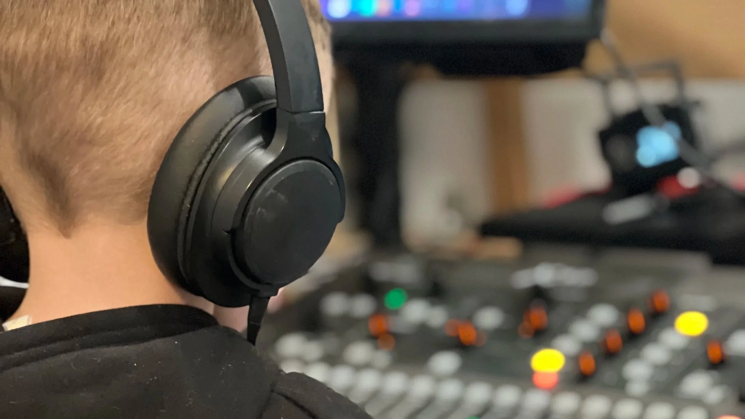 Close-up of a person with dark skin wearing black headphones near a mixing console with colorful knobs and a blurred computer screen in the background.