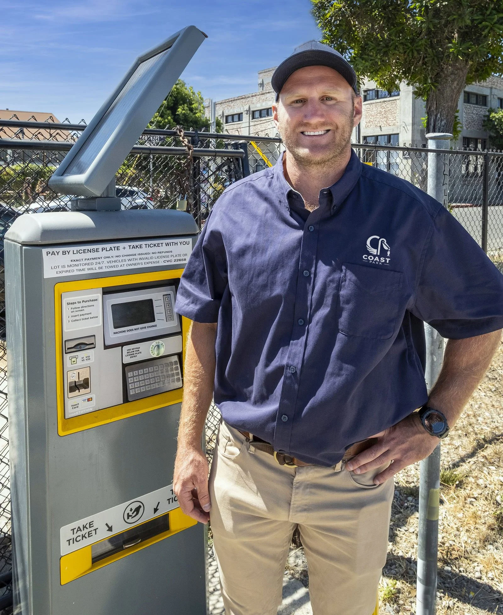 A man wearing a blue shirt and khaki pants standing next to a parking kiosk with a smile. He is outdoors, with trees, a chain-link fence, and a building in the background.