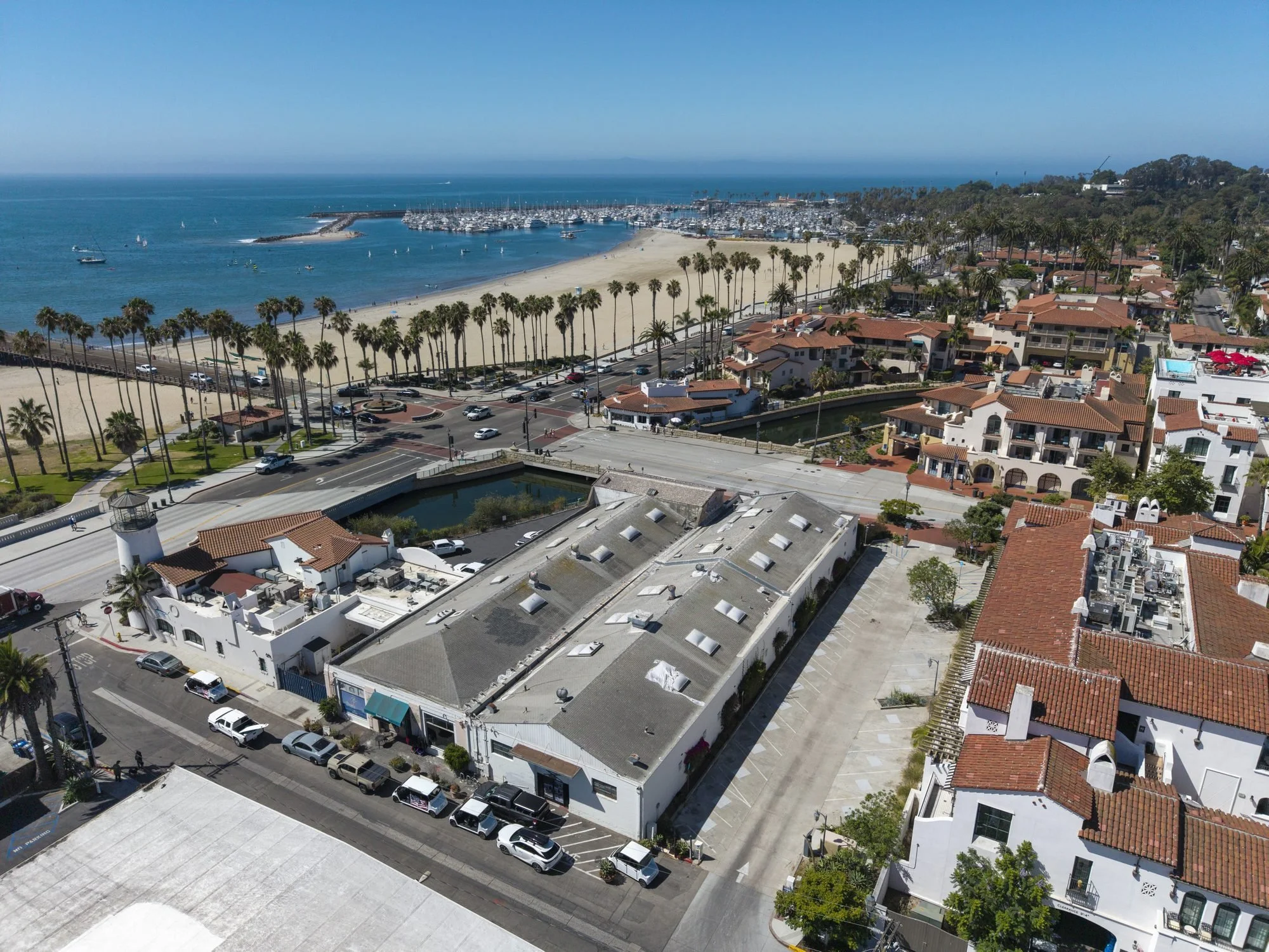 Aerial view of a beach town with a marina, sandy beach, palm trees, residential buildings with red-tiled roofs, and a parking lot.