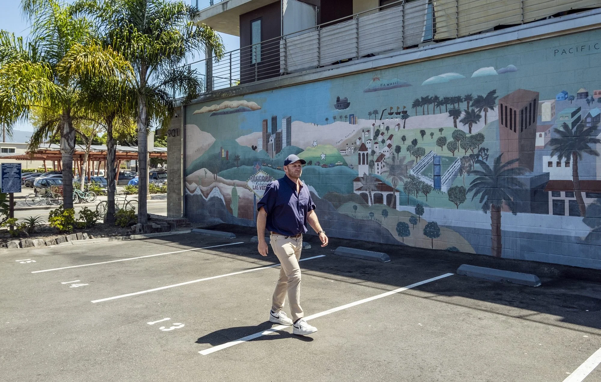 A man walking in a parking lot in front of a mural on a building wall. The mural depicts a stylized landscape with mountains, trees, buildings, and palm trees. There are palm trees and parked bicycles nearby.