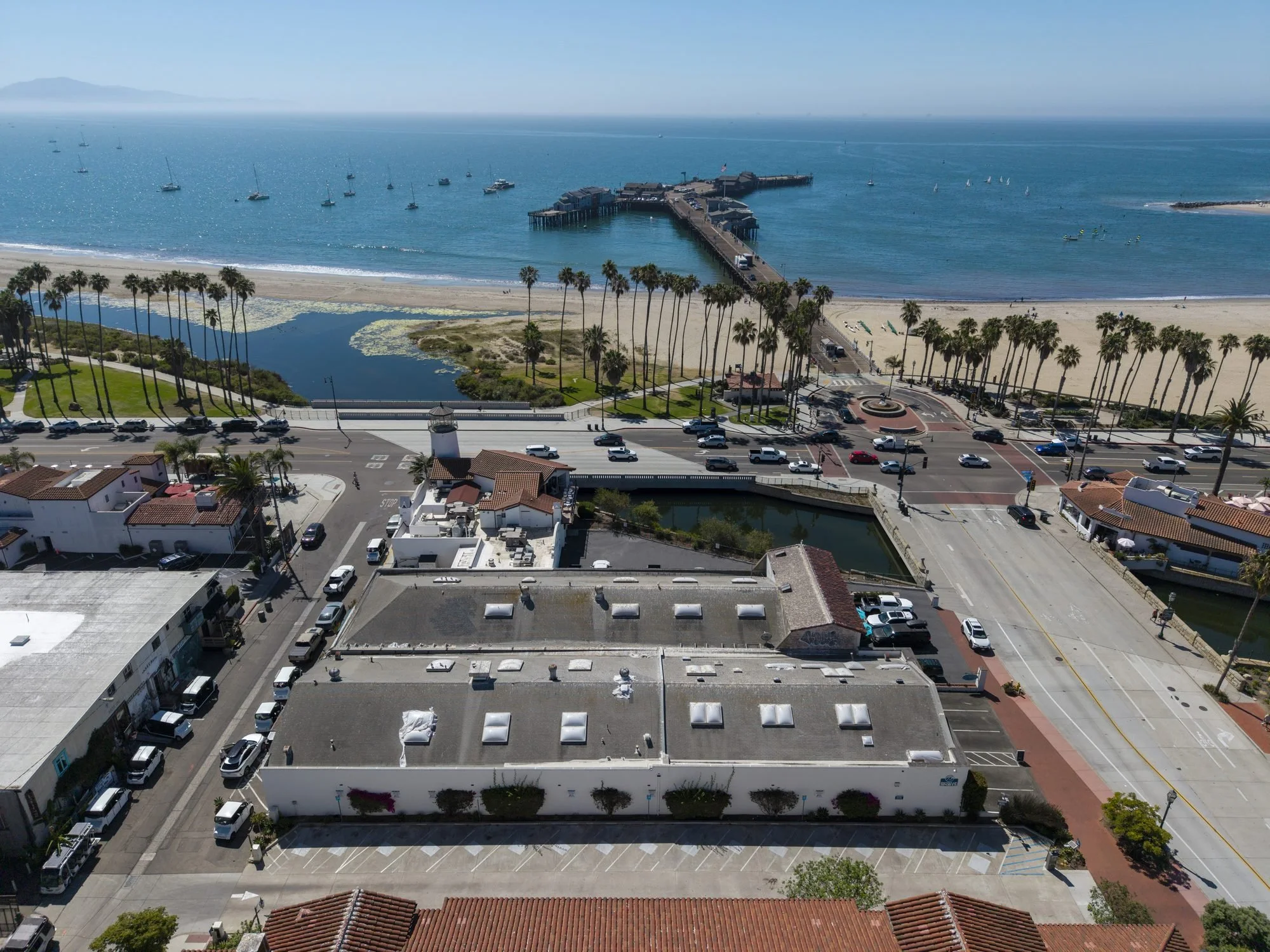 Aerial view of a beach city with a pier extending into the ocean, sailboats and paddleboards in the water, palm trees lining the sandy shore, and cars parked along the streets.