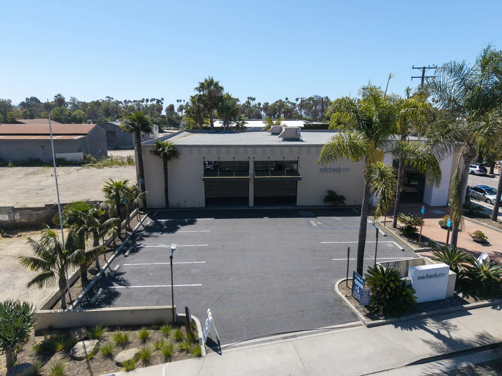empty parking lot in front of a white building labeled 'michaelkade interior design' with some palm trees and power lines, under a clear blue sky.