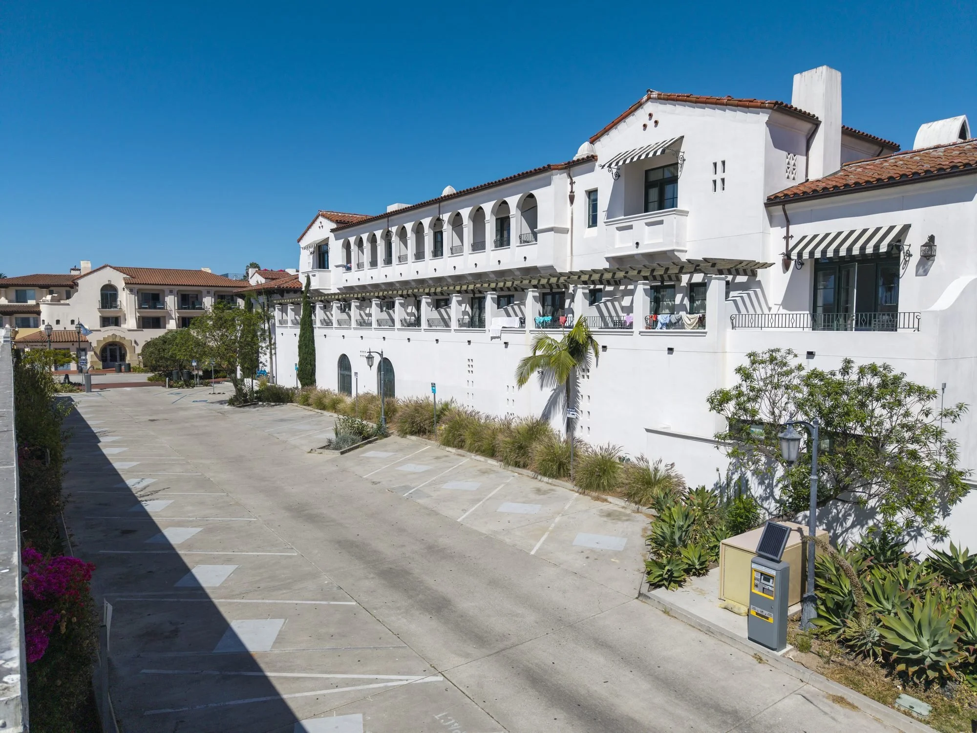 A parking lot with no cars, surrounded by white Mediterranean-style buildings with red tile roofs and balconies under a clear blue sky.