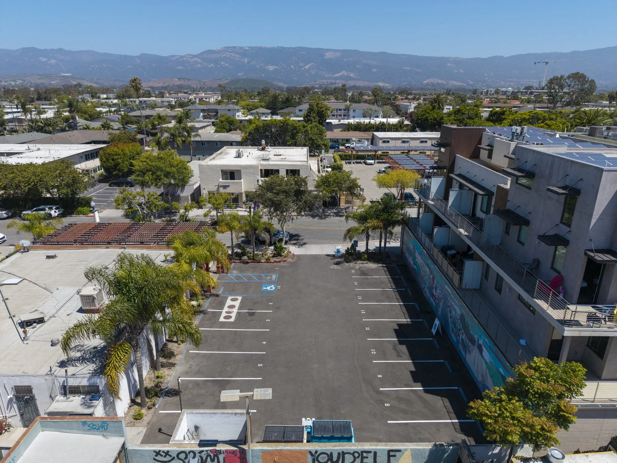 Empty parking lot with a few scattered cars, surrounded by trees and buildings, with mountains in the background under a clear blue sky.