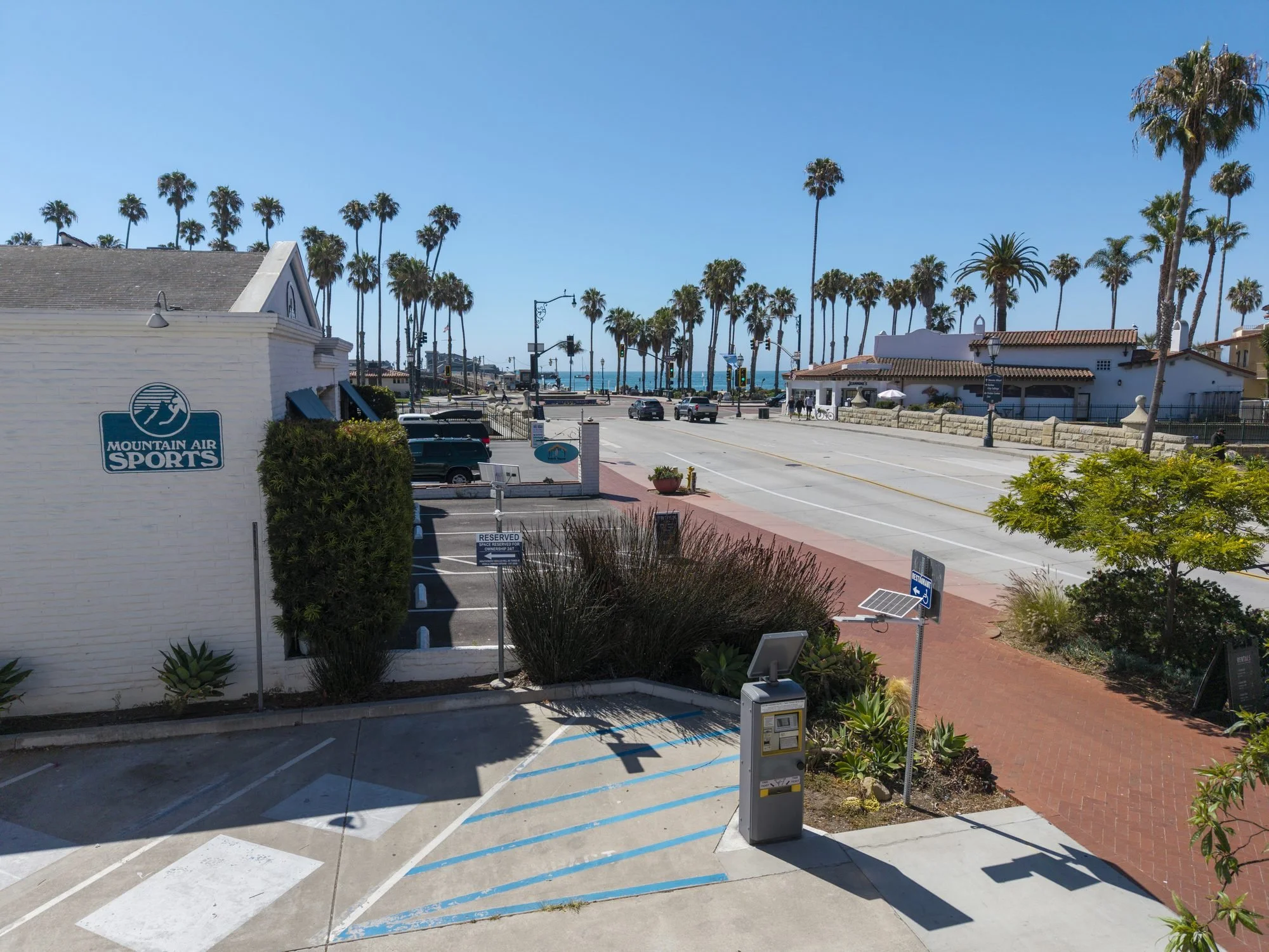 A street scene with palm trees, a beach, a white building with a Mountain Air Sports sign, parked cars, and a parking lot with a reserved sign. There are sidewalk pathways, streetlights, and sunny weather with clear blue sky.