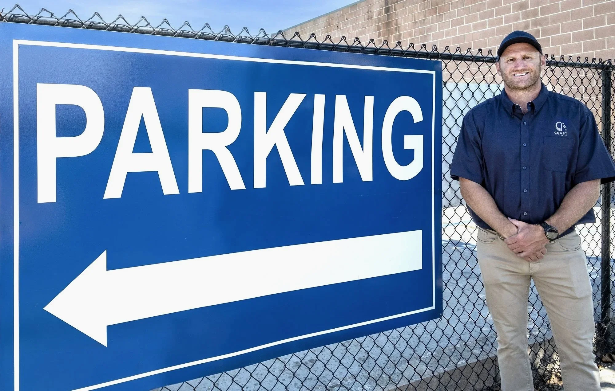 A man with a beard, wearing a navy blue cap and navy blue shirt with a logo, standing next to a large blue parking sign with a white arrow pointing left.