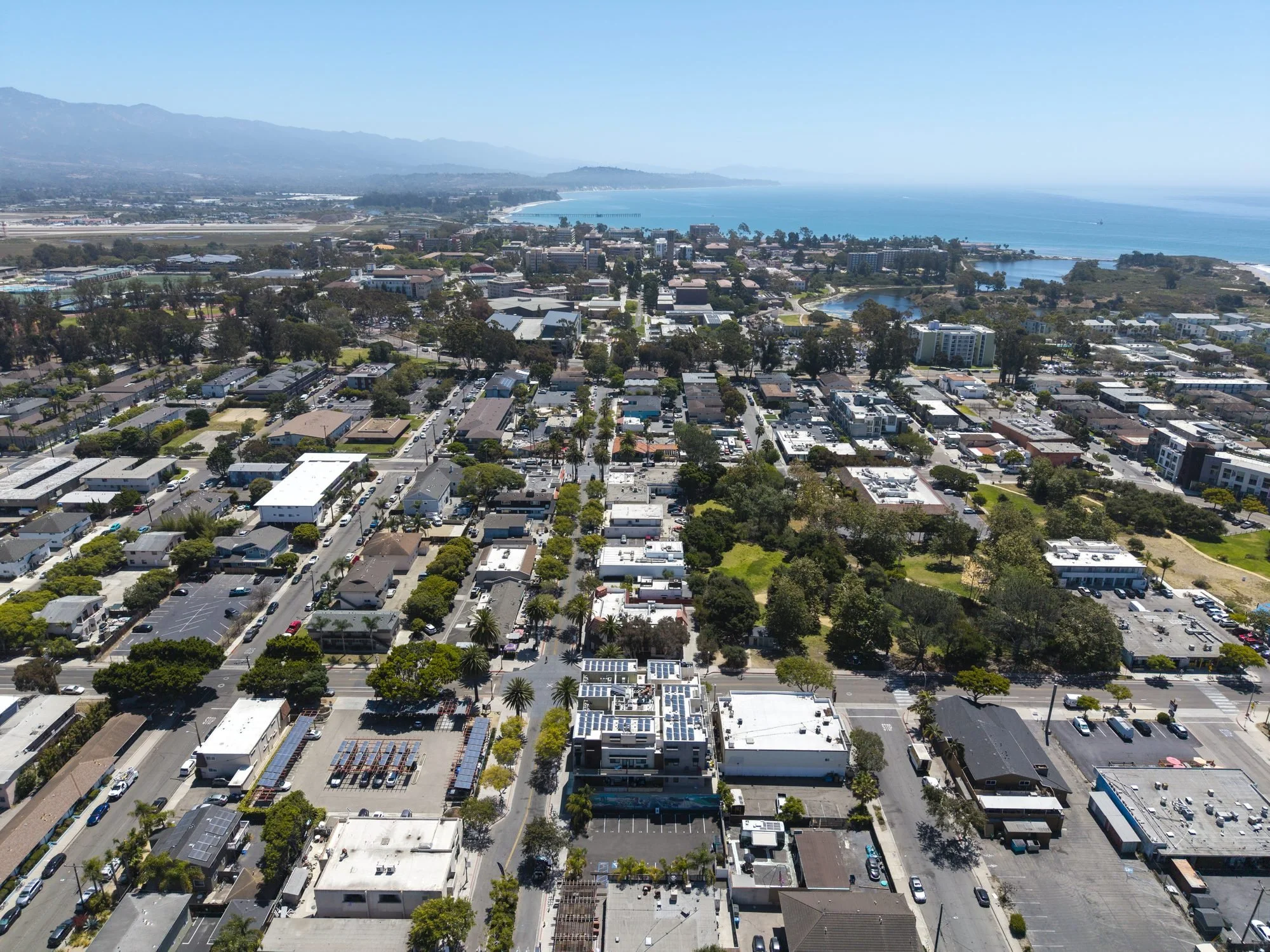 An aerial view of a coastal city with a beach along the shoreline, a bay with a small island, and a mountain range in the background. The city has residential and commercial buildings with some trees and parking lots.