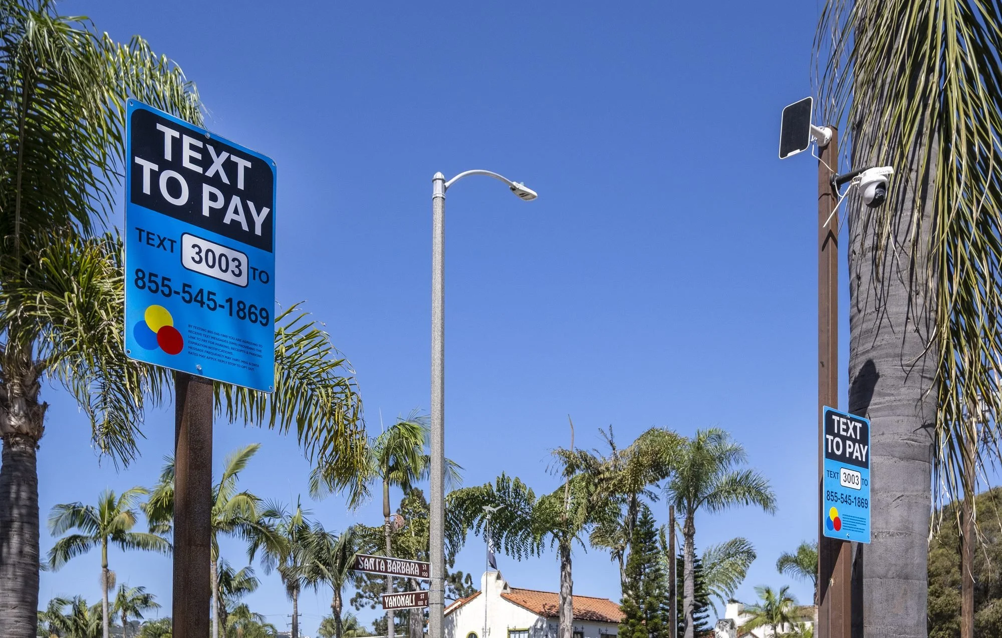 Street scene with palm trees, street signs, a blue sky, and a pole with a surveillance camera and a solar panel.