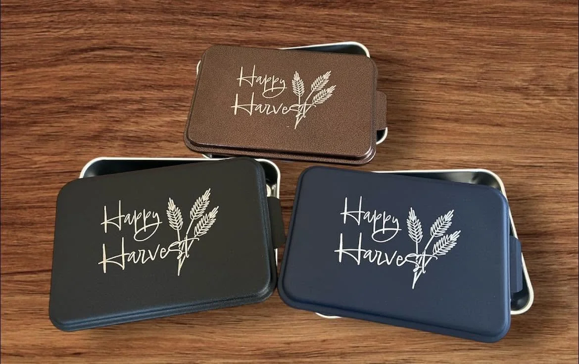Three rectangular tins with a harvest theme, labeled "Happy Harvest" and decorated with wheat illustrations, placed on a wooden surface.