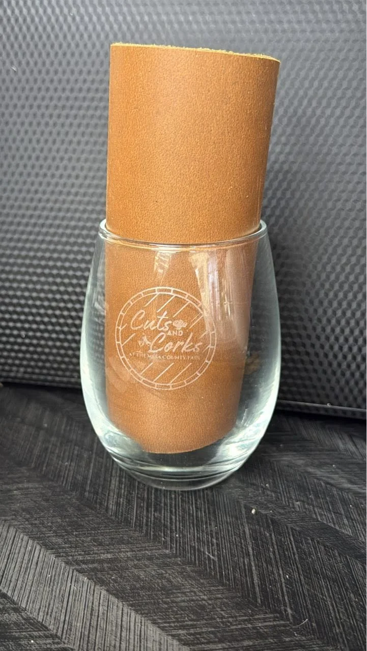A clear glass with the logo 'Cuts and Curls at the top, the column' printed on it, holding a rolled piece of brown craft paper, placed on a dark wooden surface with a textured dark background.