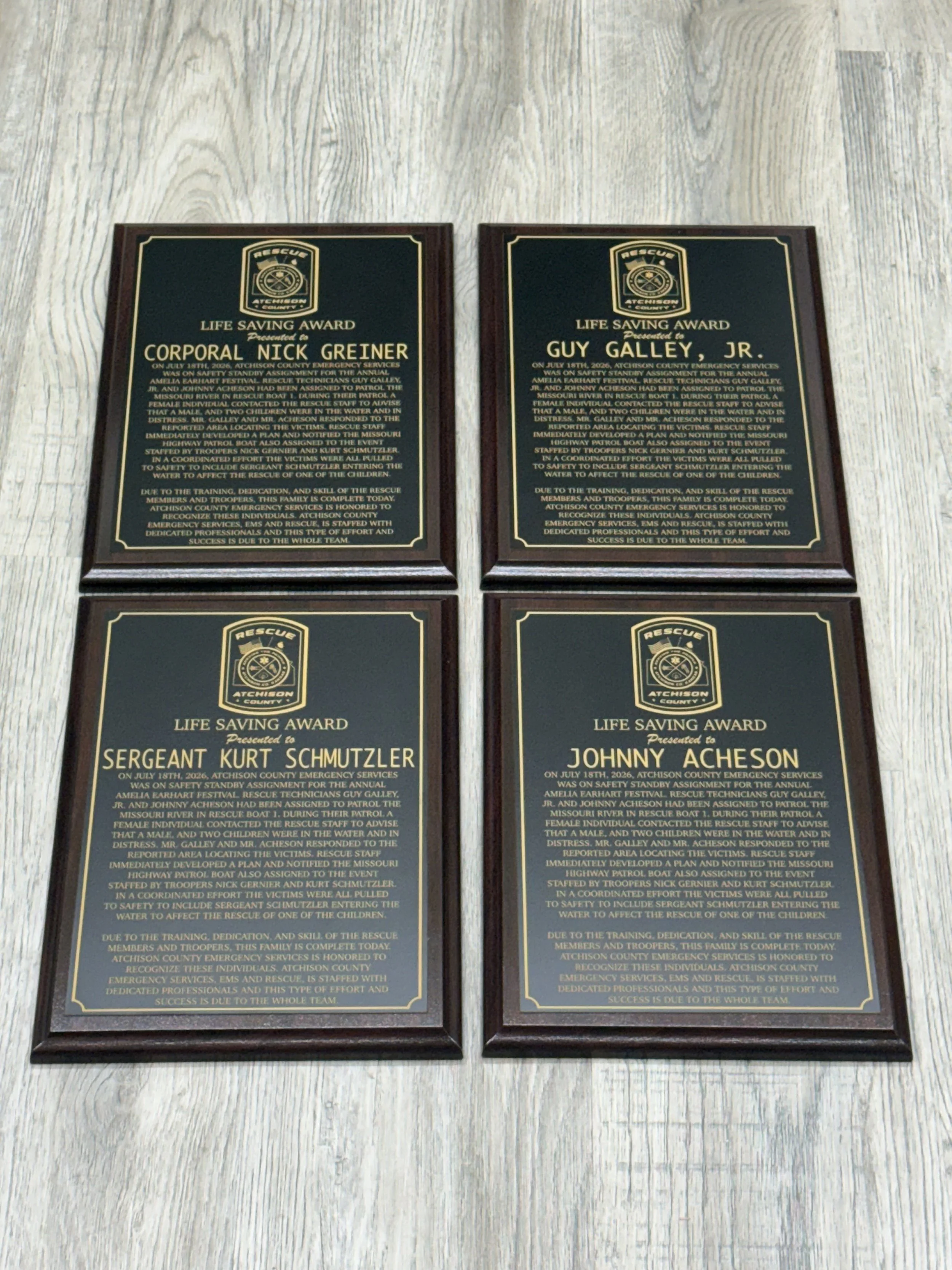 Four wooden plaques with black and gold text, each recognizing a different individual for a life-saving award from Atchison County emergency services, arranged on a light-colored wooden surface.