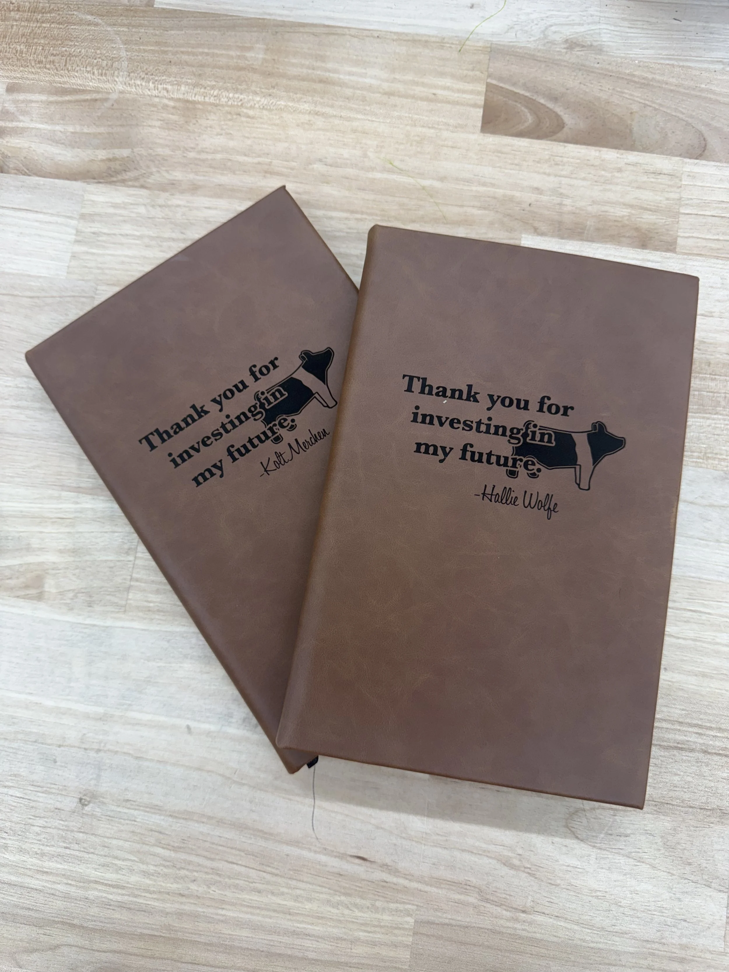 Two brown notebooks with motivational quotes about investing in the future, placed on a light wooden surface. One notebook is partially on top of the other.