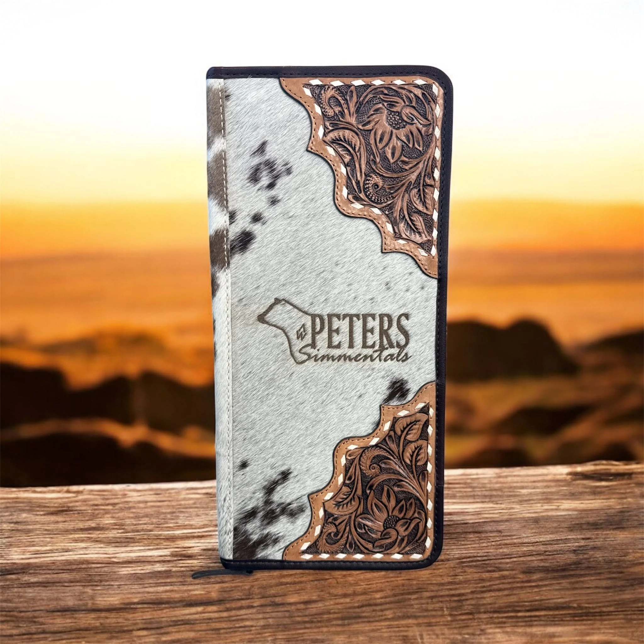 Leather wallet with intricate floral carving on one side and cowhide pattern on the other, placed on a wooden surface with sunset background.
